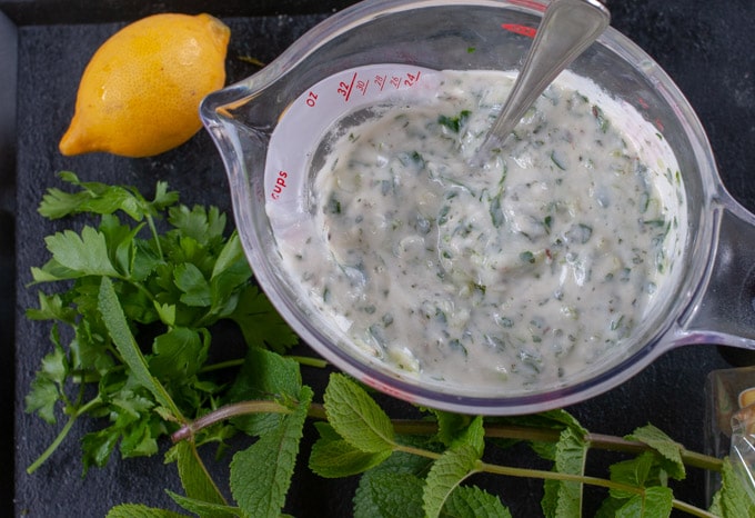 yoghurt sauce in a bowl with a spoon and herbs and lemon on the side of the bowl