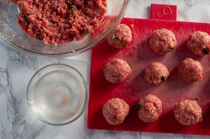 meatball mixture in a bowl, some meatballs on a cutting board and a small bowl with warm water 
