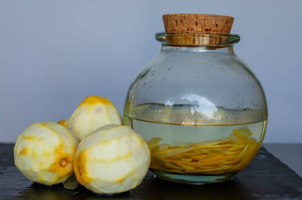 lemon zests infusing in alcohol inside a jar and peeled lemons on the side